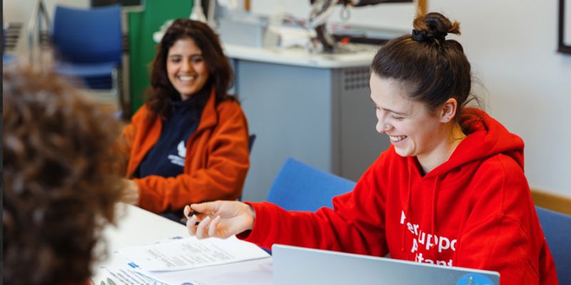 A group of students sitting around a table in a classroom, smiling and chatting.