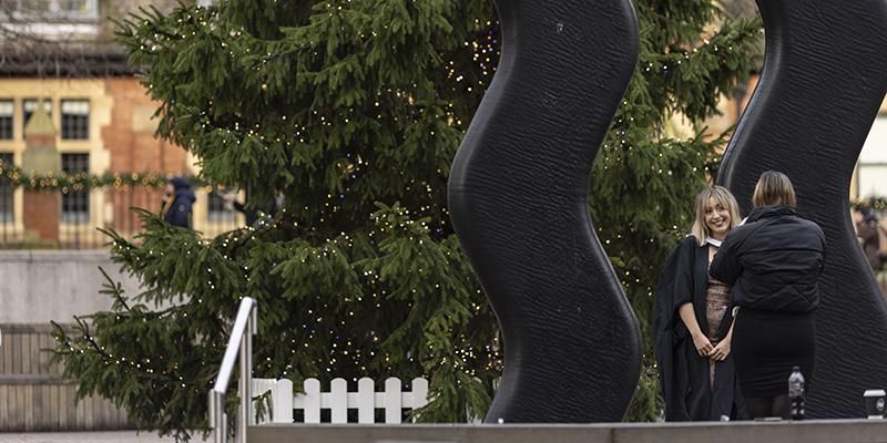 Graduate having their photo taken in front of the Sign for Art sculpture and a Christmas tree