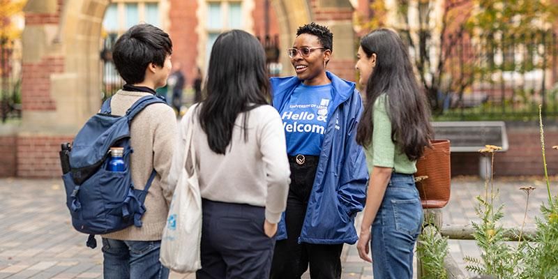 An International welcome team ambassador wearing a blue top talking to three students on campus