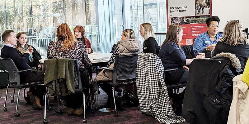 About ten adults are seated at two round tables, chatting. Coats hang on chair backs. Floor-to-ceiling windows and a glass building are visible outside. A red noticeboard includes the message “It’s never too late to learn.