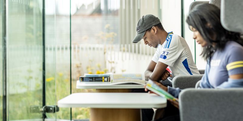 Two students sit in a bright campus study area near floor-to-ceiling glass windows, reading books at small tables.