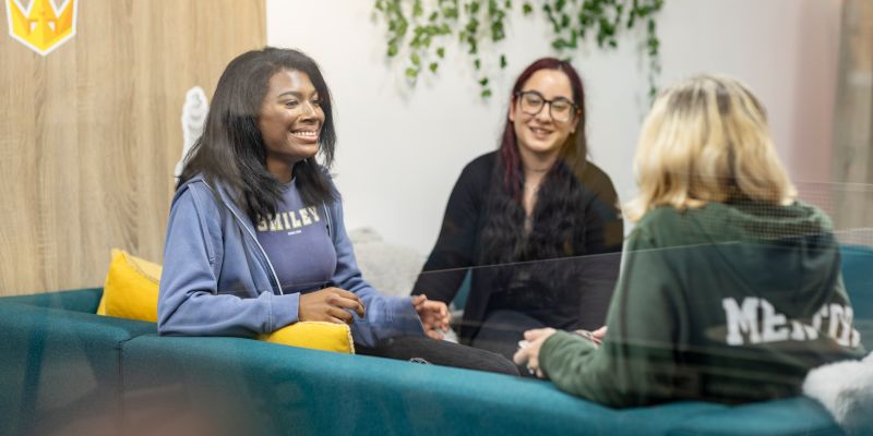 Three students sit together in a comfortable campus lounge, engaged in a supportive conversation.