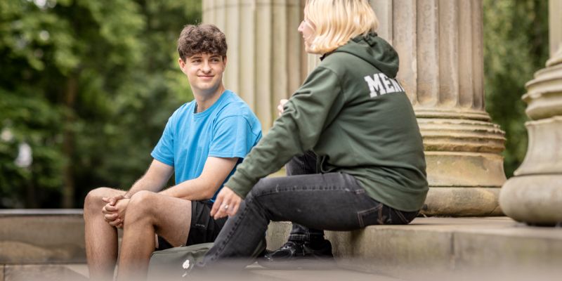 Two people sitting on the stone steps of a historic building with large columns, talking together.