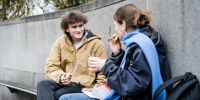 Two students sitting together chatting. One is eating a sandwich