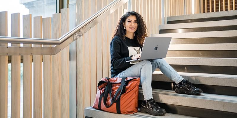 A student sitting on some steps with a laptop on their knees and a large orange bag next to them