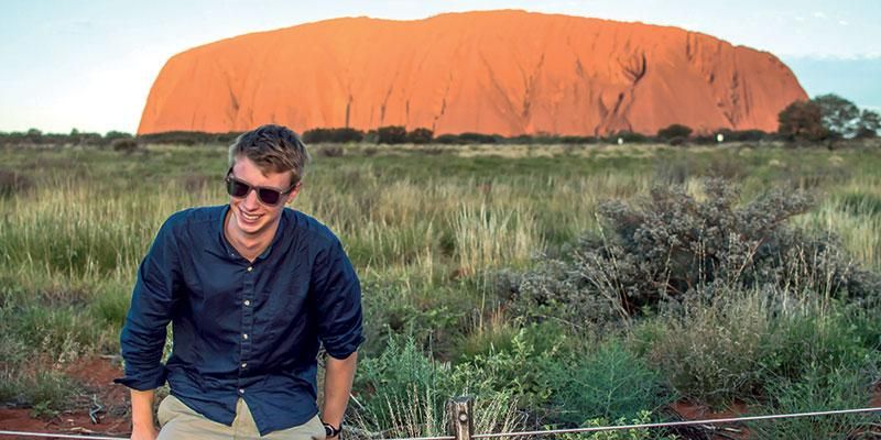 A student wearing sunglasses is standing in front of Ayers Rock in Australia.