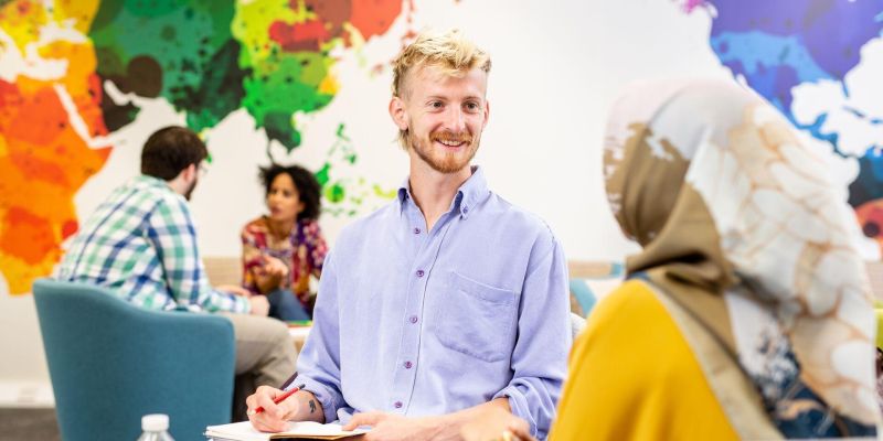 A postgraduate research student sits inside a large communal space, which has a colourful abstract world map on the wall and chats to another person while writing in a notebook.