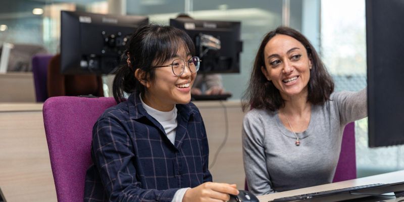 A tutor providing technical support to a student whilst sat at a desk working on a computer