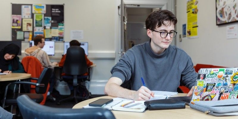 A students sits at a round table in a teaching space. They are looking at an open laptop, with a pen poised to write on a notebook in their hand. There are people working, chatting and using computers in the background.