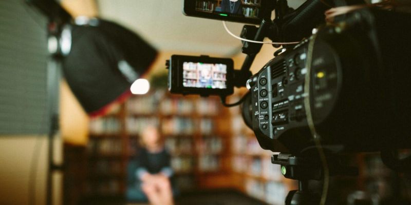 A camera in the foreground points at a person sat in a chair in the background
