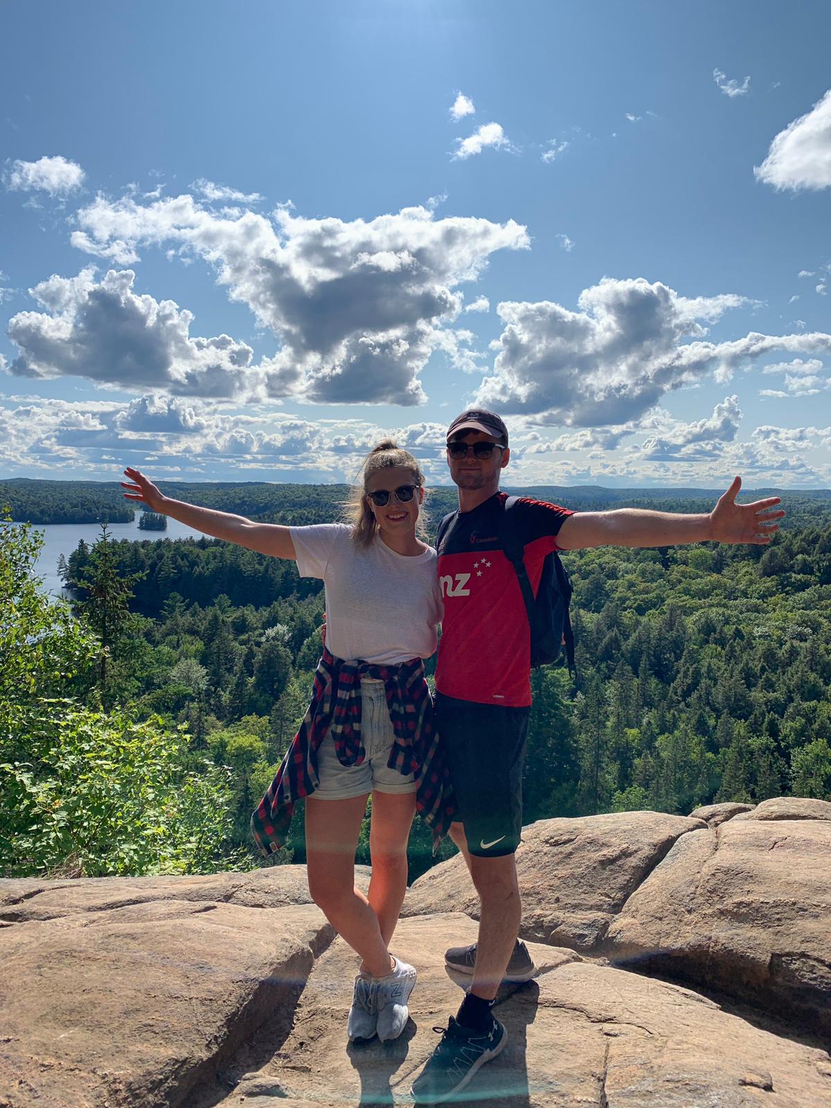 Two people stand with their arms outstretched on top of rocks overlooking a forest and water