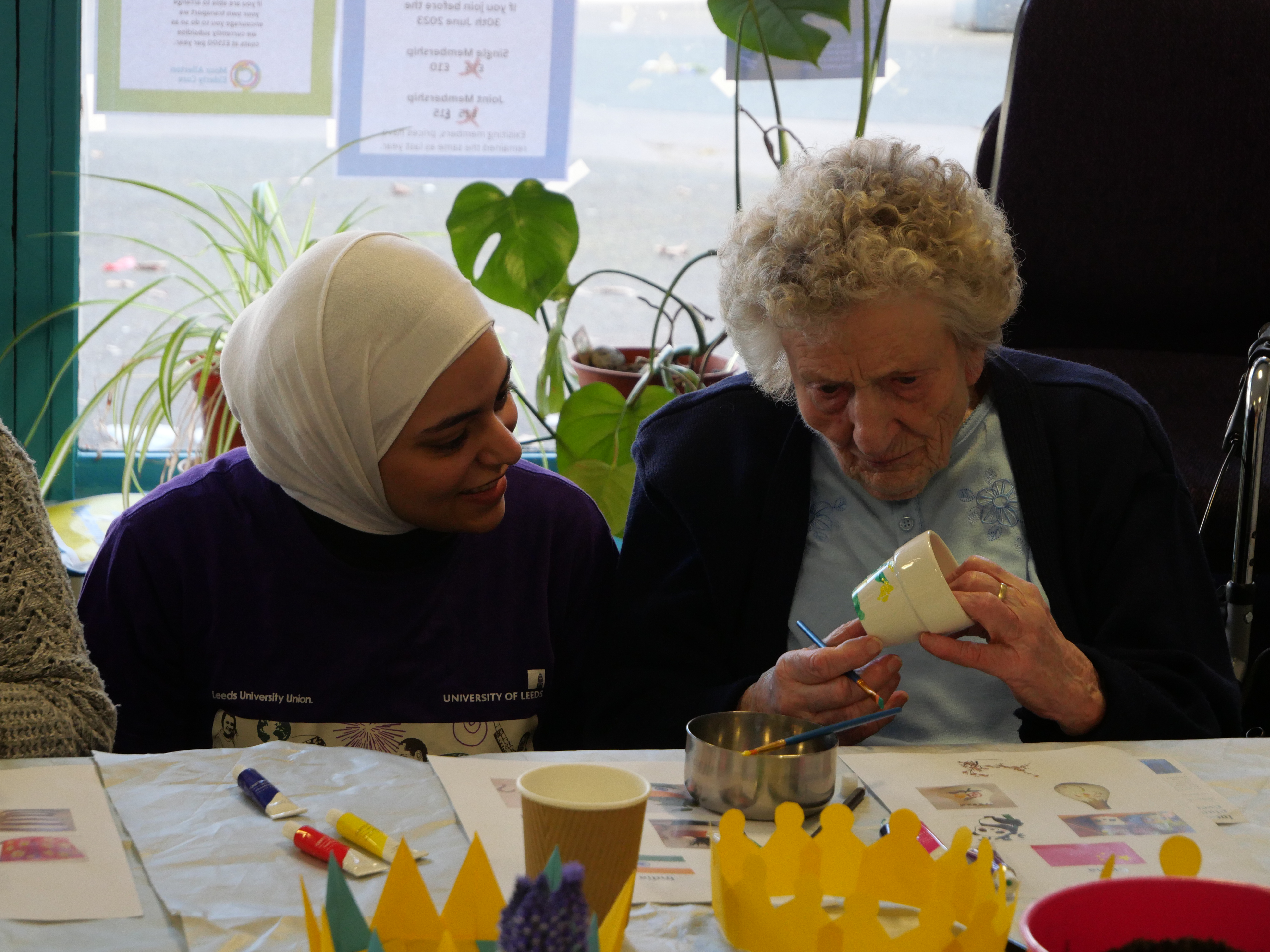 A student wearing a hijab talking to an elderly resident while she's painting a plant pot.