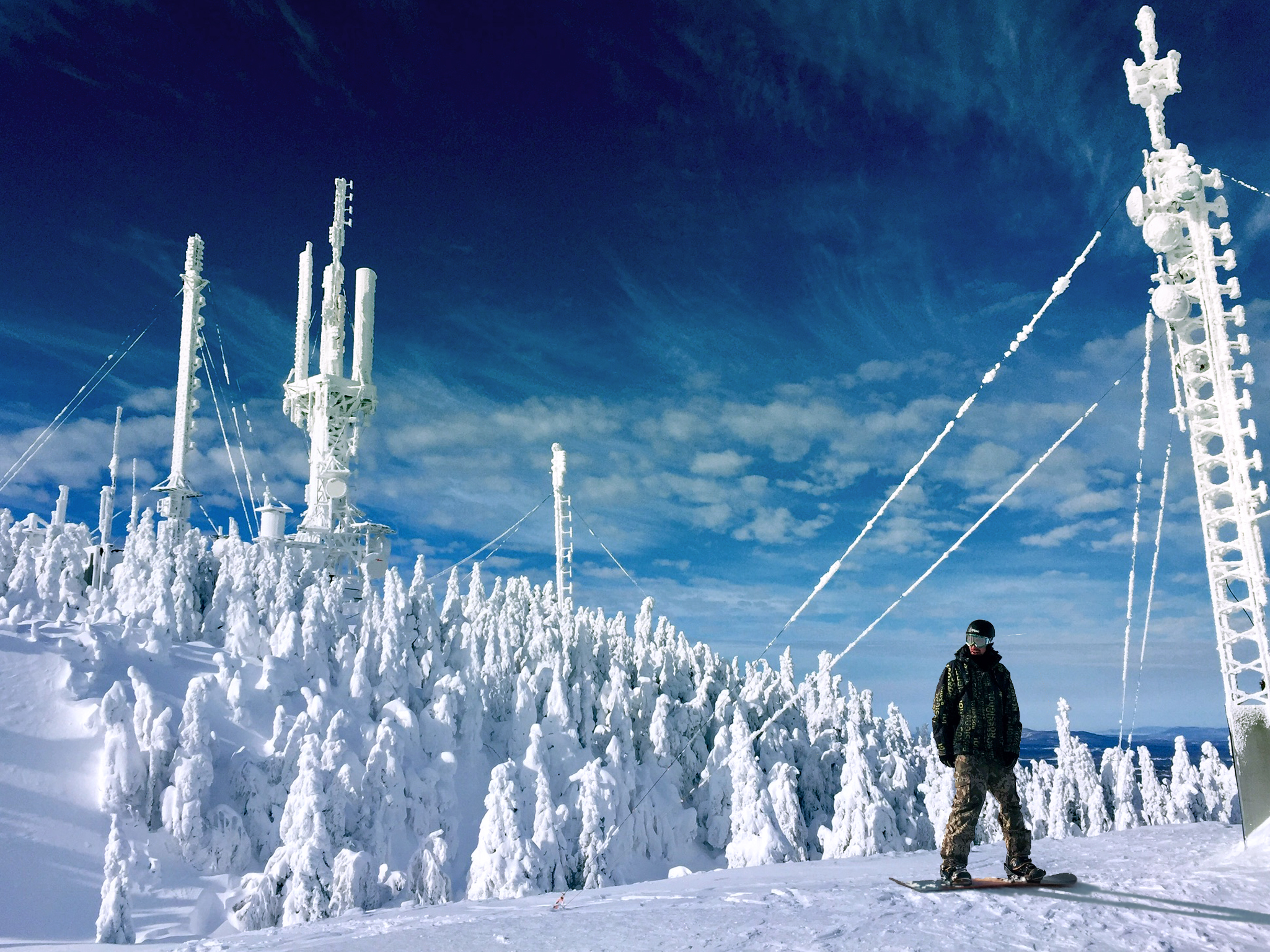  A person dressed in ski gear,  who is stationary on a snowboard in a snowy area. There are  trees covered in snow in the background and the sky is blue with some cloud. 