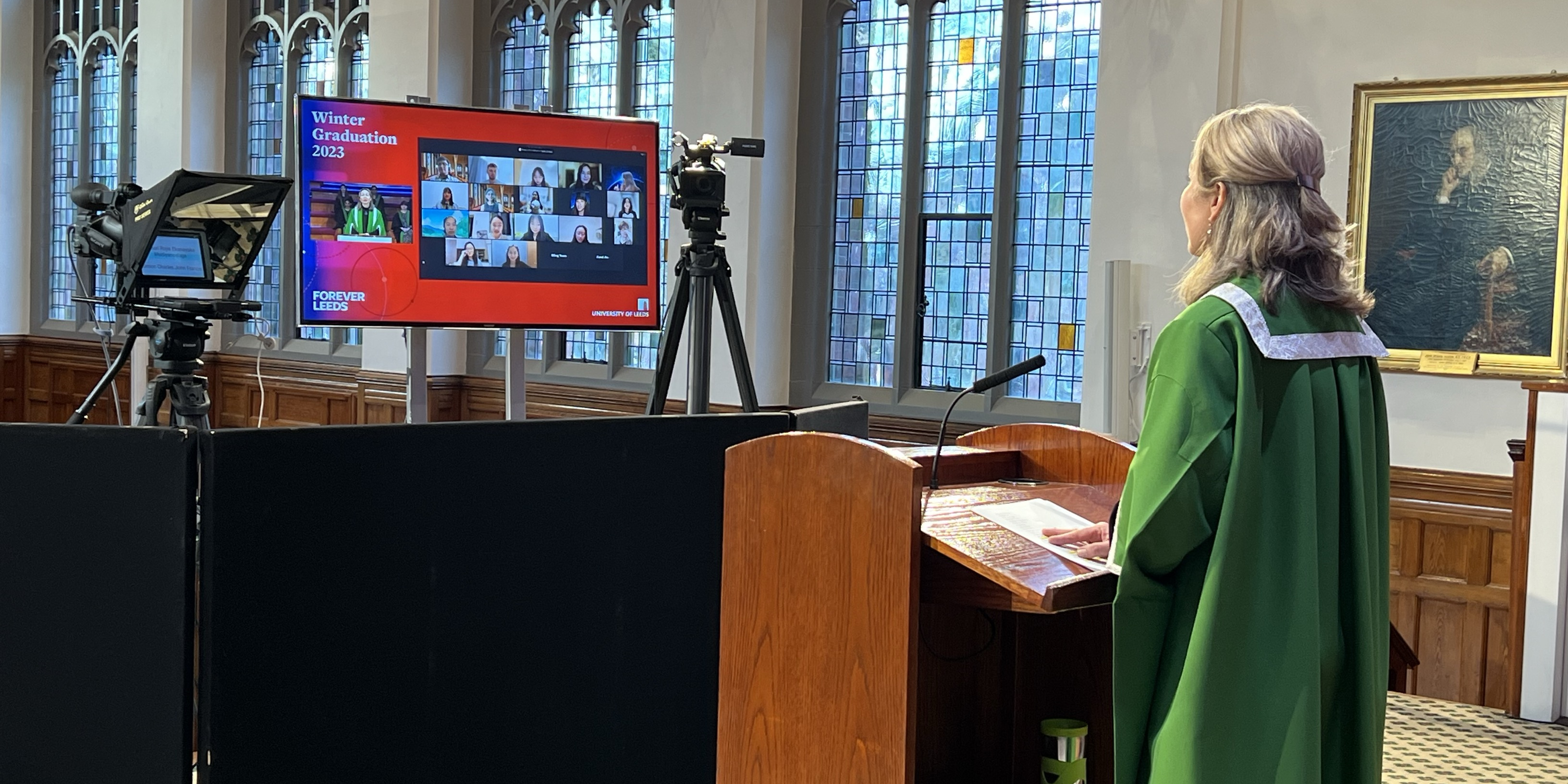 A presenter stands in a formal green robe at a podium. She looks at a large screen with the faces of virtual graduates looked back. 