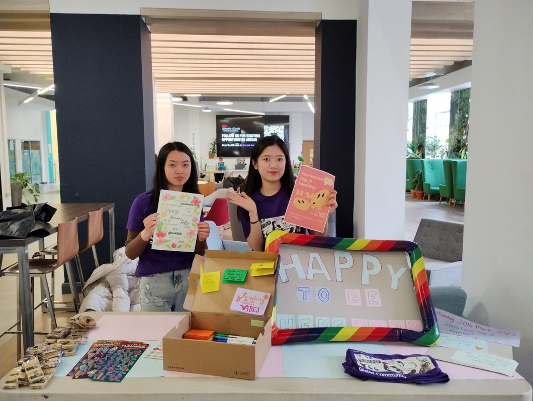 Two students holding signs behind a table in the LUU. Stickers, pens and paper on the table for students passing by to leave comments and messages. 